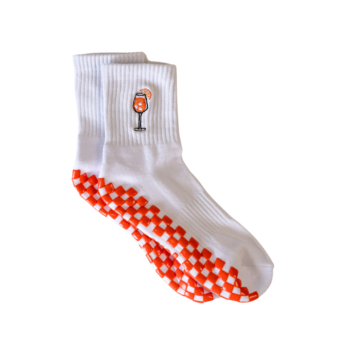 The Spritz Grip Sock by Revel: white socks with a small Aperol Spritz cocktail embroidery and bold orange-and-white checkerboard grips on the soles—perfect as Pilates grip socks. Plain white background.
