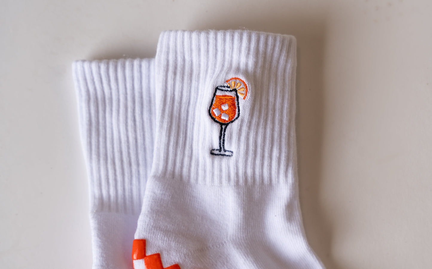 The Spritz Grip Sock by Revel: white ribbed socks, one embroidered with a wine glass holding an orange drink and slice. They’re displayed on a white surface with one sock overlapping the other.