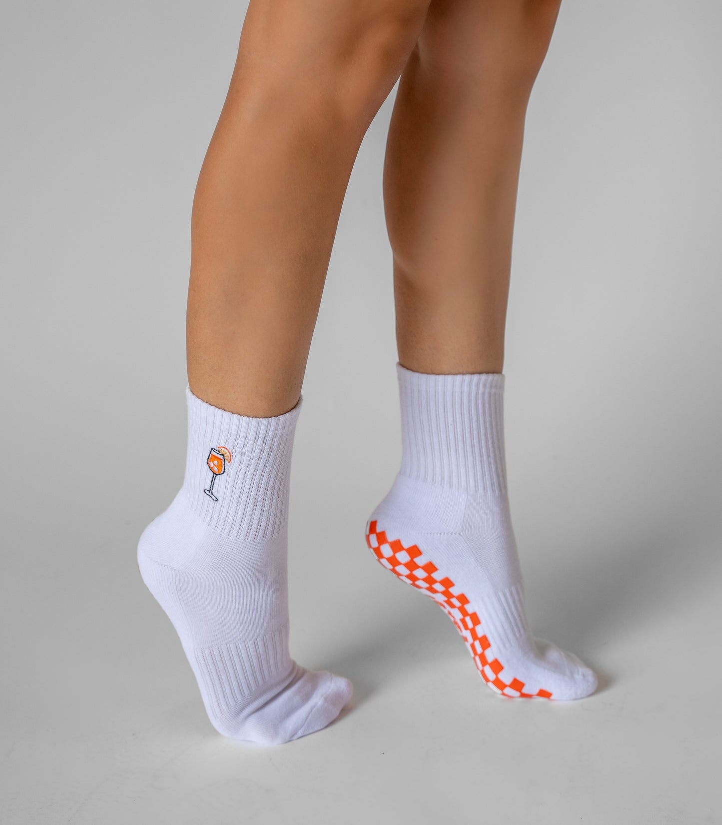 A person stands on tiptoe in white Revel The Spritz Grip Socks, featuring a small orange wine glass graphic on the left ankle and orange checkerboard grip soles—ideal as playful Pilates grip socks. The backdrop is plain light gray.