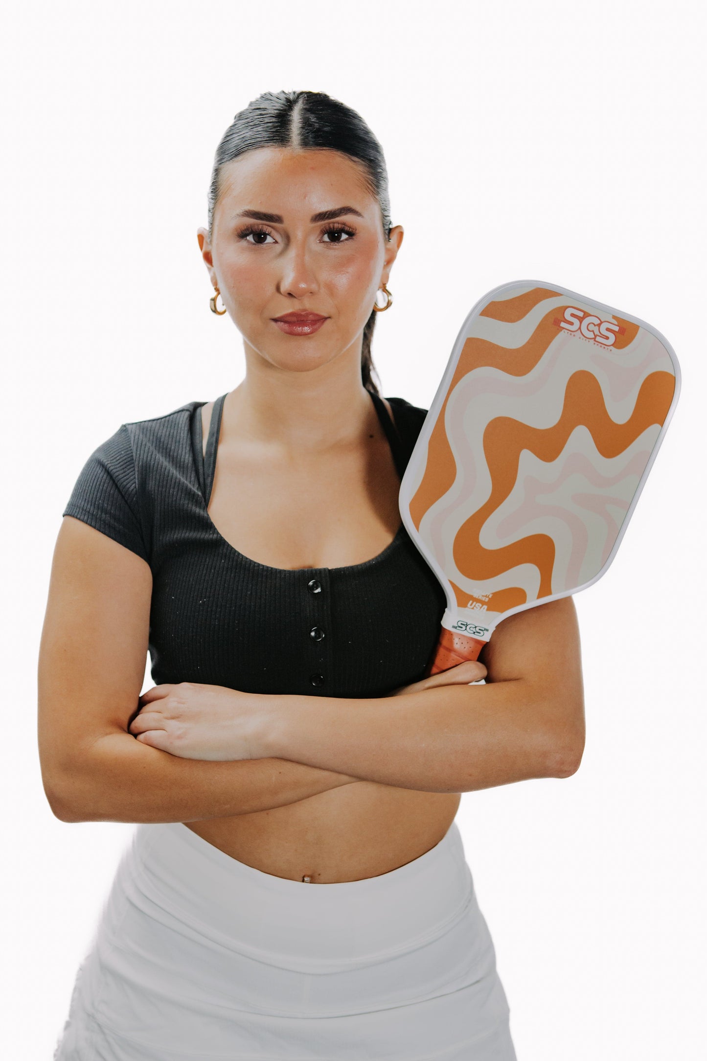 A confident woman with a sleek ponytail holds The Winston Evo-Lite Series paddle by Salted City Sports, featuring an orange and beige wavy design. She sports a black cropped top, white skirt, and gold hoops, ready to play with her USA Pickleball approved paddle.