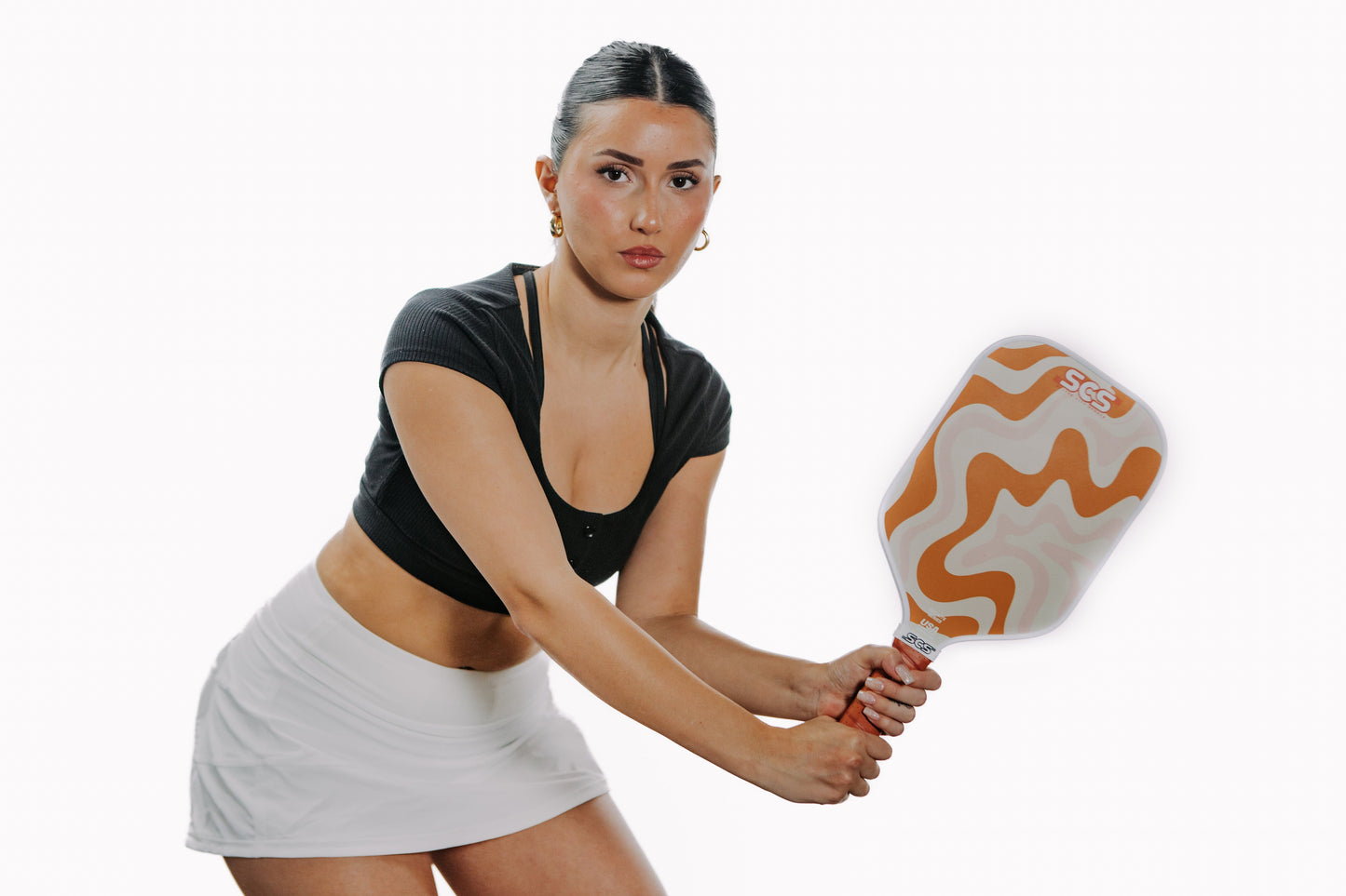 A woman with dark hair tied back wears a black crop top and white tennis skirt, holding The Winston | Evo-Lite Series pickleball paddle by Salted City Sports (USA Pickleball approved) in an athletic stance against a white background.