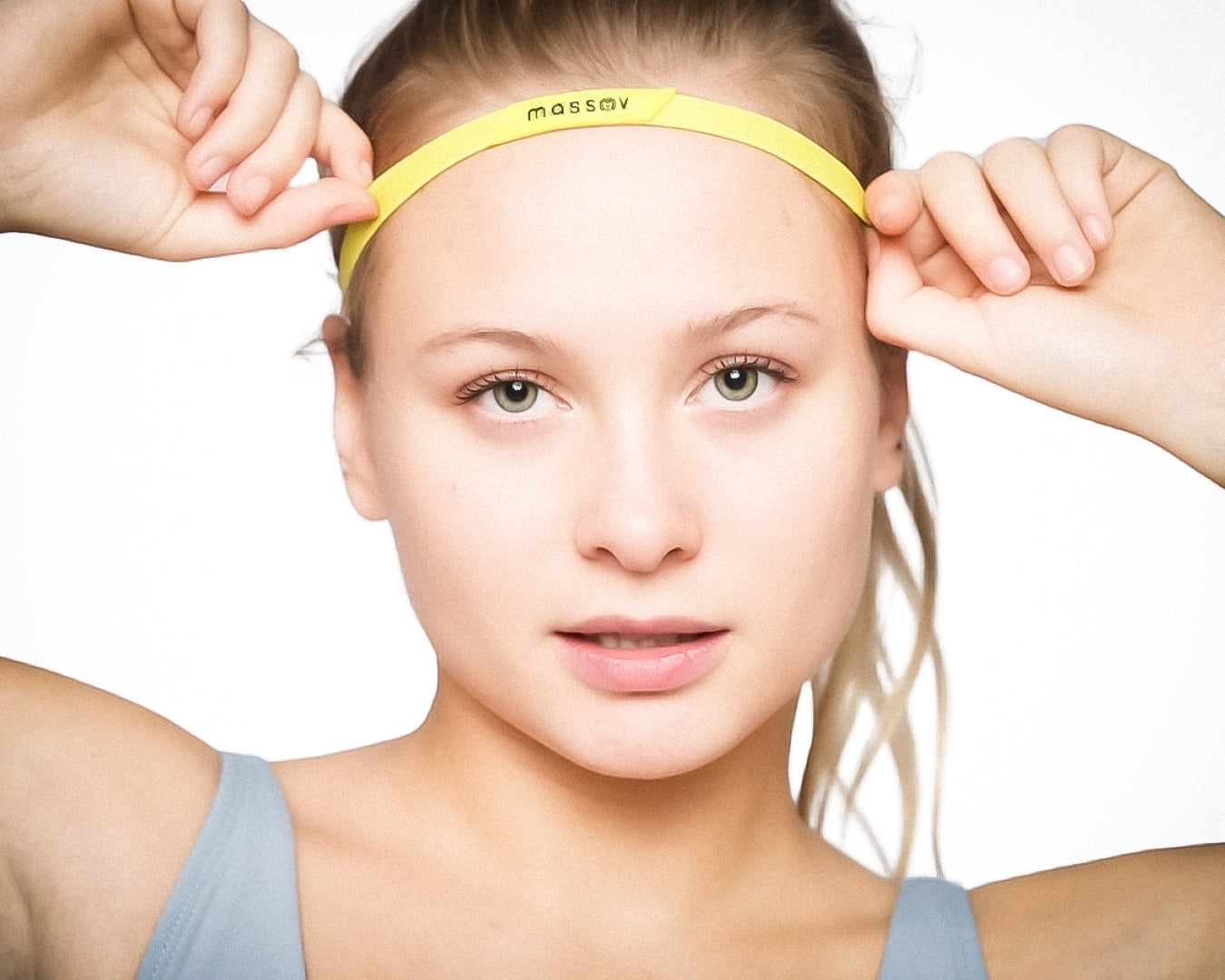A young woman with fair skin and light hair wears a light blue top and adjusts a yellow Massov Thin Sports Headband with both hands, posing against a plain white background.