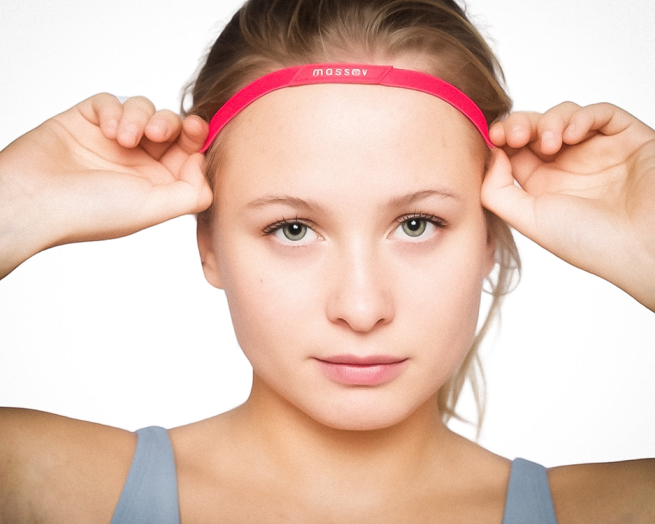 A young person with fair skin and light eyes wears a pink Massov Thin Sports Headband on their forehead, paired with a light blue tank top. Their light brown hair is pulled back as they face the camera against a plain white background.