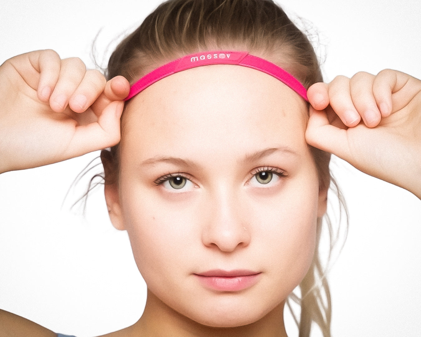 A fair-skinned young woman with light eyes looks at the camera while adjusting a bright pink Massov Thin Sports Headband. Her hair is pulled back, her expression is neutral, and she uses both hands to hold the headband.