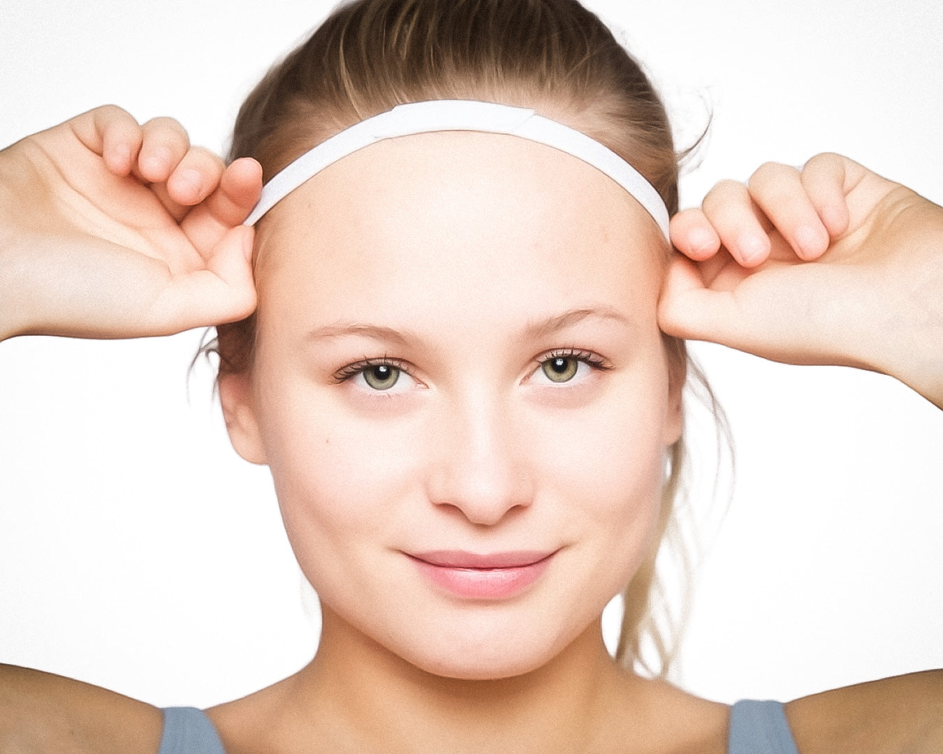 A young woman with fair skin and light eyes smiles gently as she adjusts the Massov Thin Sports Headband on her head. She faces the camera in a light blue tank top against a clean, minimal background.