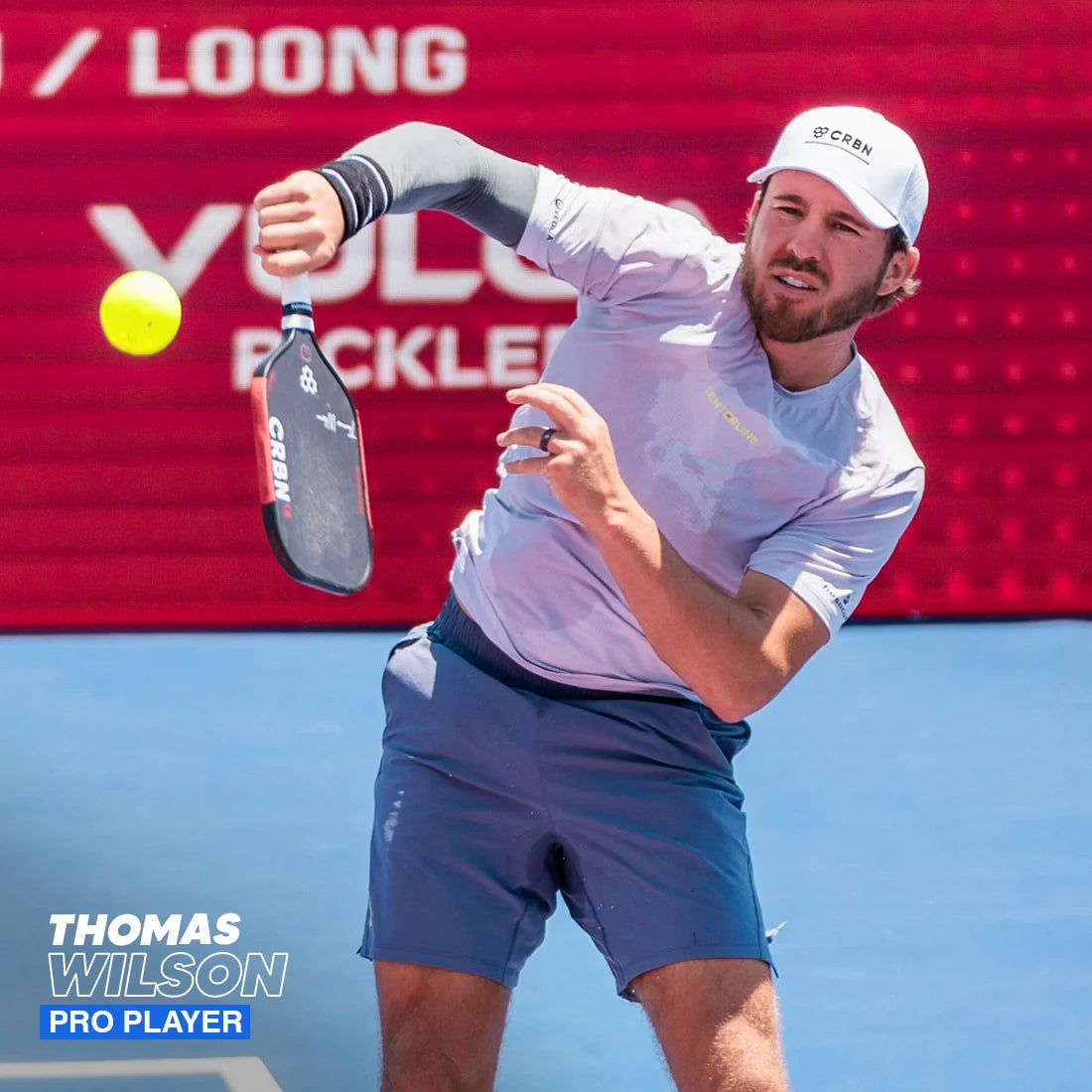 A male pickleball player in a light grey shirt, blue shorts, and white hat swings his paddle—fitted with a Hesacore Grip Pickleball Elongated Grip (6” long, regular feel, white)—at a yellow ball on a blue court. Red background with “Thomas Wilson, Pro Player” below.