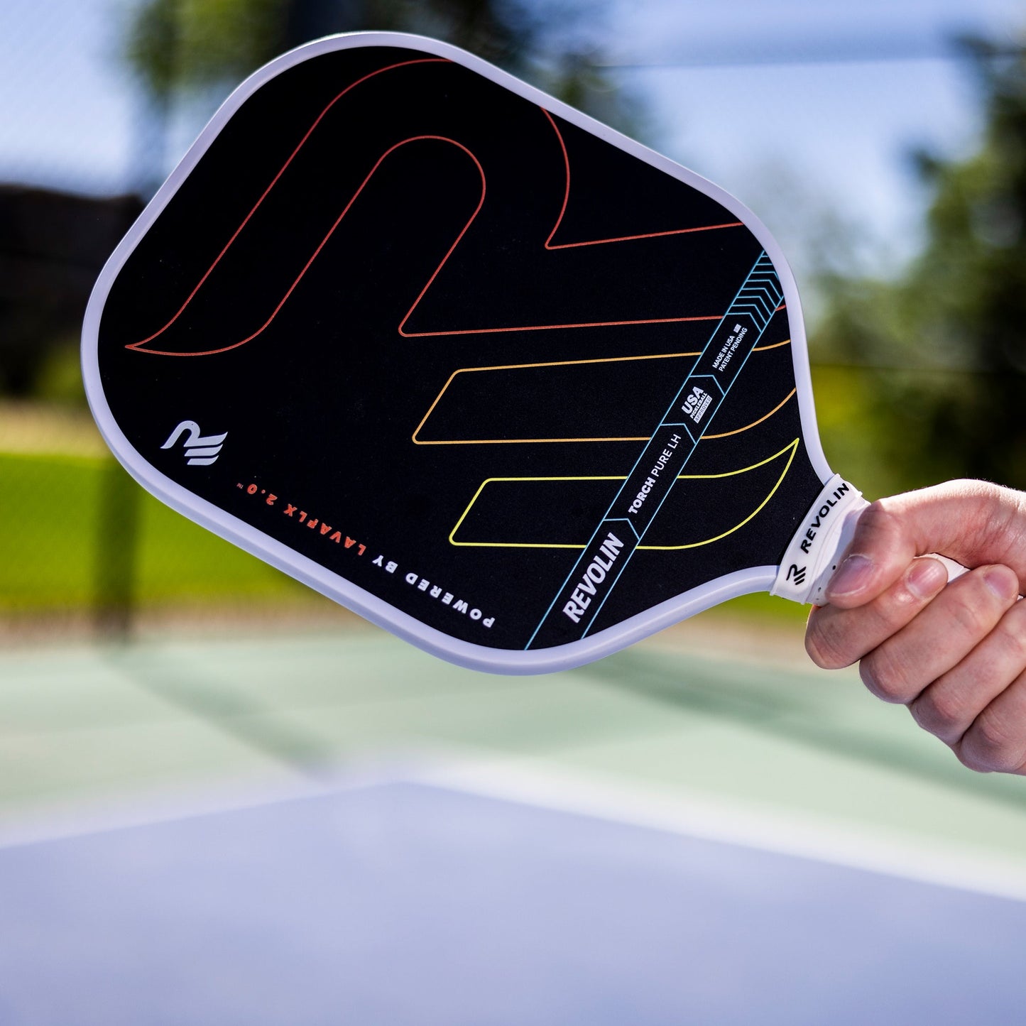 A person holds a Revolin Sports Torch Pure LH Pickleball Paddle with colorful curved lines and text at an outdoor court, featuring a blurred green background and blue surface below, all brightly lit by sunlight.
