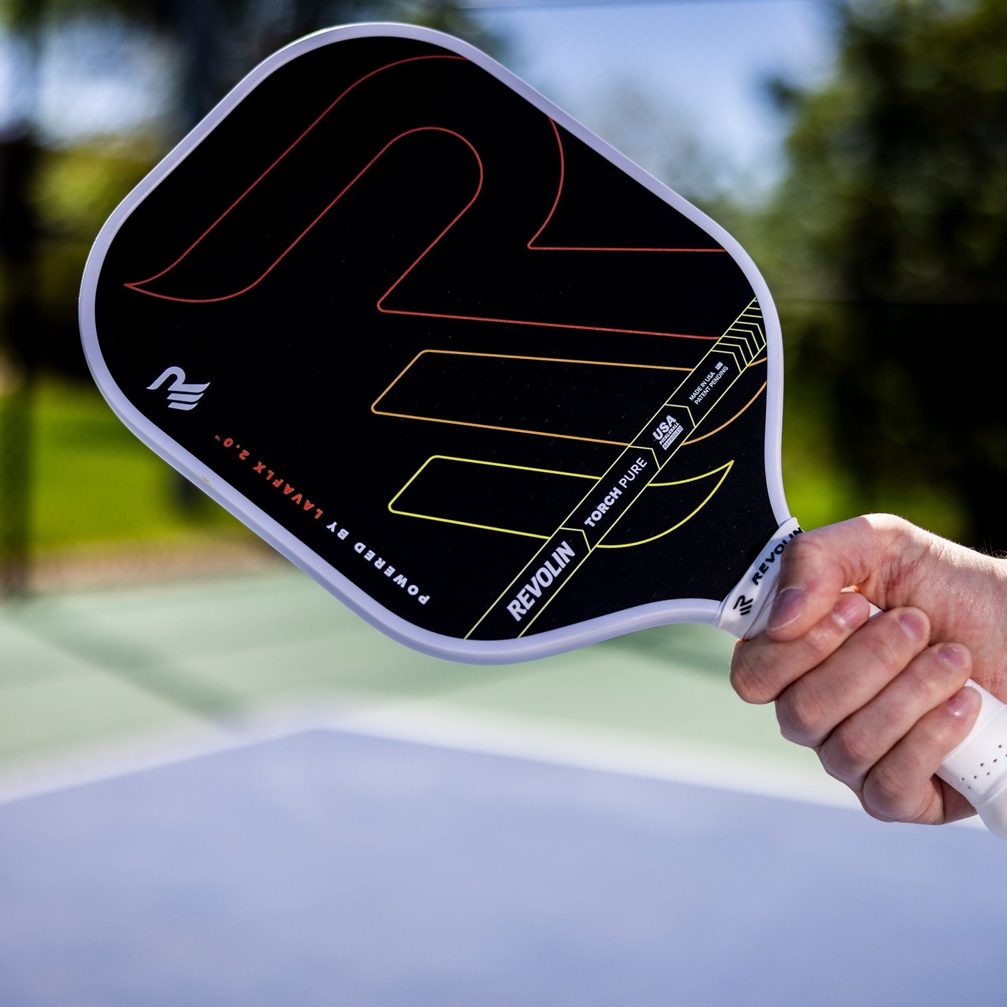 A close-up of a hand holding the black Revolin Sports Torch Pure Pickleball Paddle with red, orange, and yellow accents. The paddle features a polymer honeycomb core and is shown on an outdoor court with greenery and clear sky.