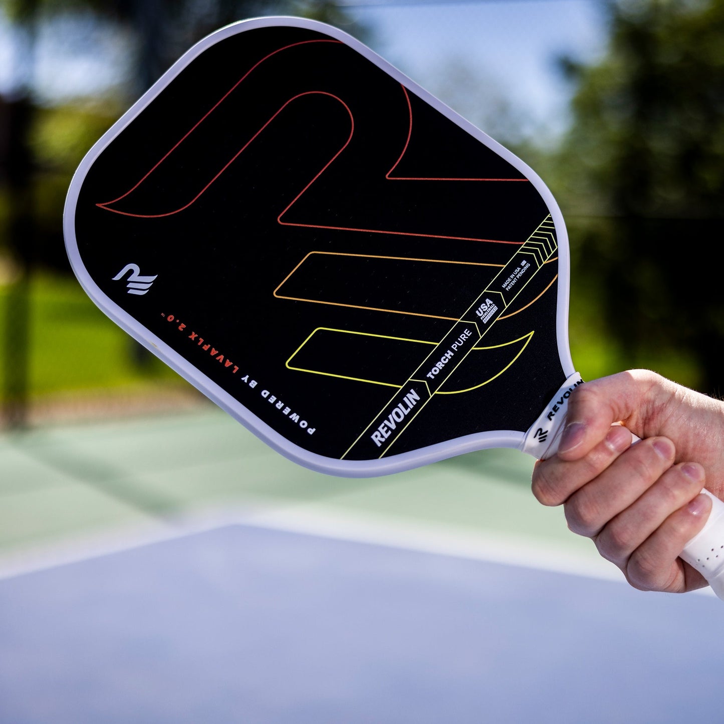 A hand holds the Revolin Sports Torch Reach Pickleball Paddle, featuring bold red and yellow lines, a 3D-printed octagonal grip, and "REVON" branding; an outdoor court and greenery blur in the background.