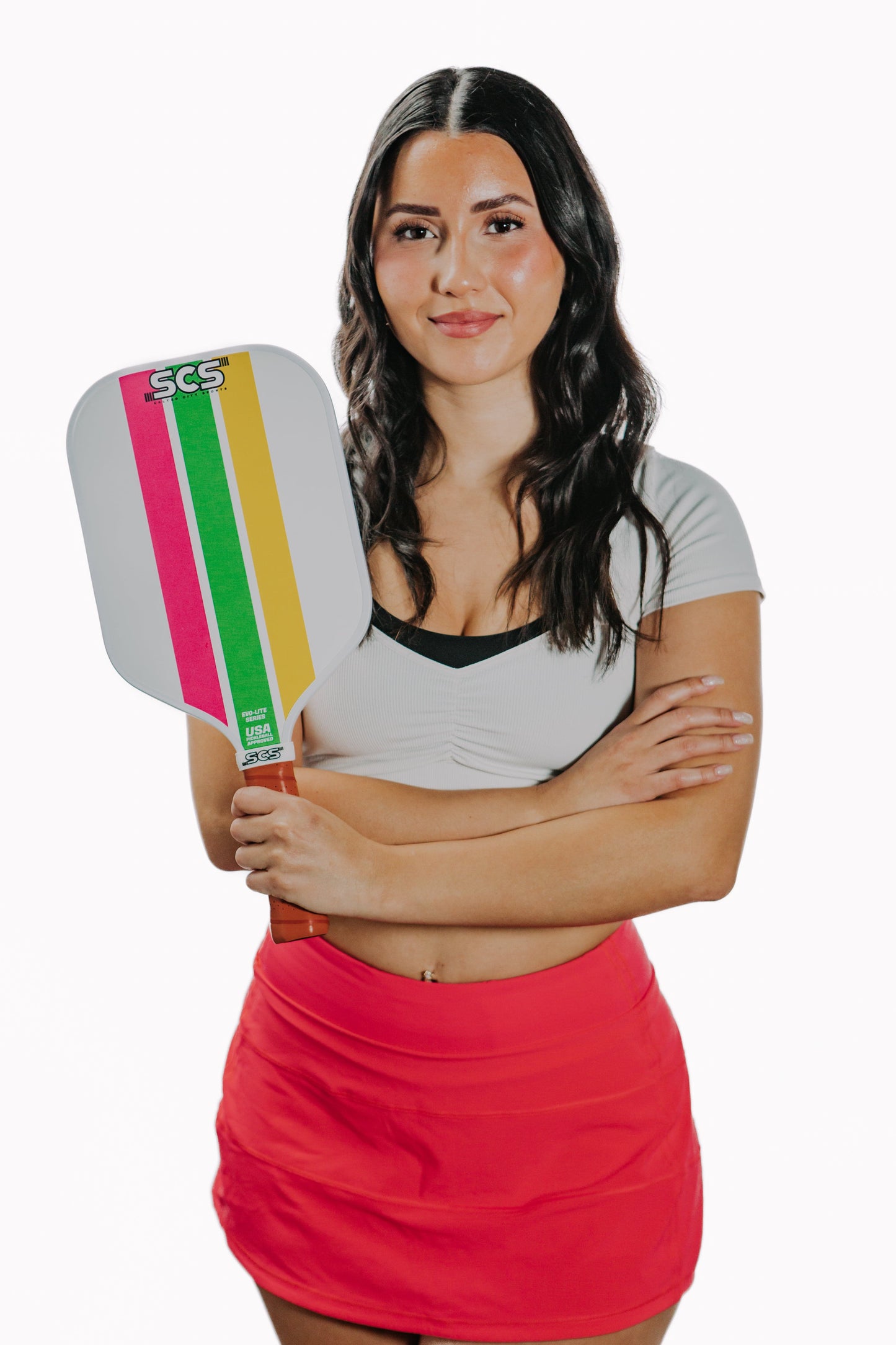 A woman with long dark hair stands confidently, arms crossed, holding the Salted City Sports Trifecta | Evo-Lite Series USA Pickleball approved paddle with multicolored stripes. She wears a white crop top and red athletic skirt against a plain white background.