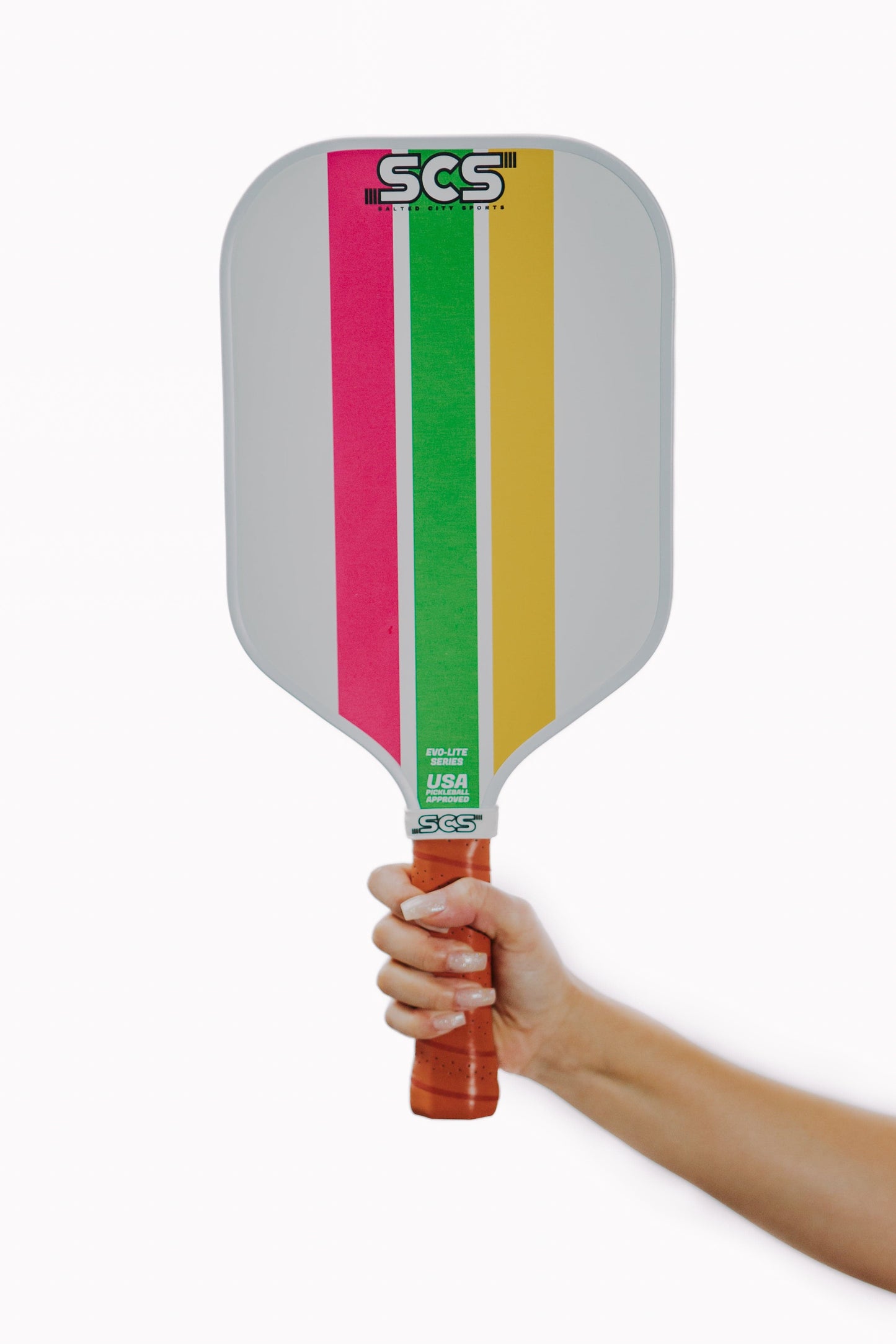 A hand holds the Salted City Sports Trifecta | Evo-Lite Series pickleball paddle, featuring a brown leather grip, a light gray face with pink, green, and yellow stripes, the SCS logo, “USA” on top, and USA Pickleball approval. White background.