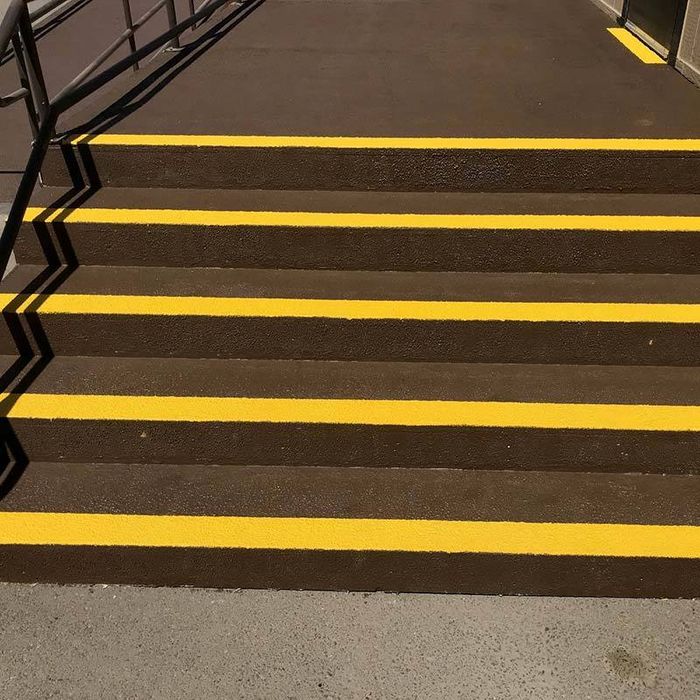 Five brown concrete steps with straight yellow edges are coated in SlipDoctors Tuff Grip Extreme Aggressive Traction Non-Skid Floor Paint. A metal handrail is on the left, and the areas above and below the stairs are flat and smooth.