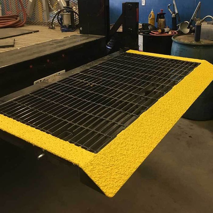 A metal grated ramp features SlipDoctors Tuff Grip or Tuff Grip Extreme, an aggressive traction non-skid floor paint with a textured yellow anti-slip coating on the edges for visibility and safety in an industrial workshop setting.