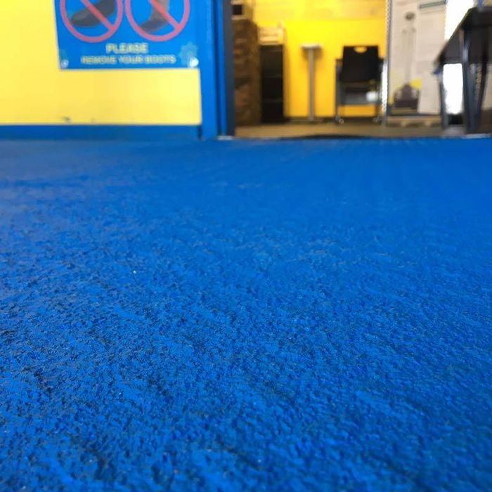 Close-up of a bright blue SlipDoctors Tuff Grip or Tuff Grip Extreme Aggressive Traction Non-Skid Floor Paint, featuring a vivid color and rough texture in a room with yellow walls; blurred furniture and an instruction sign appear in the background.