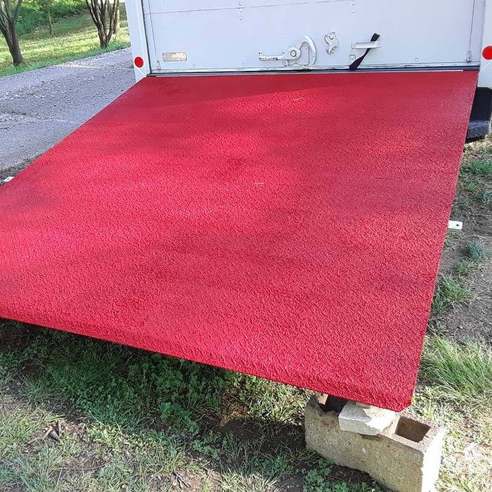 A truck’s loading ramp, covered in bright red carpet and coated with SlipDoctors Tuff Grip Extreme Aggressive Traction Non-Skid Floor Paint, extends from the vehicle onto cinder blocks over grass and dirt, with some pavement visible in the background.