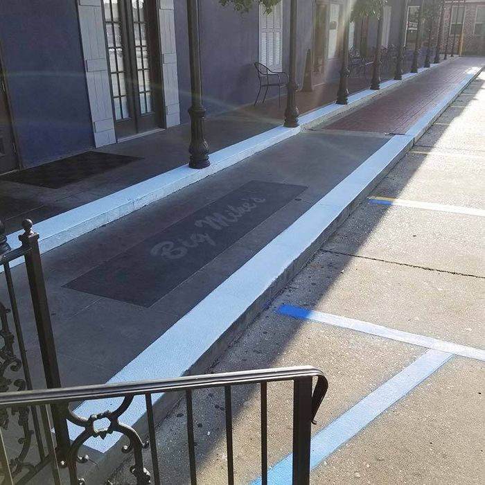 A sidewalk edged with white-painted curbs and SlipDoctors’ Tuff Grip Extreme Non-Skid Floor Paint offers high traction around a dark area labeled “Big Mike’s.” Black lamp posts, a brick wall, windows, a metal bench, and blue-marked parking are nearby.