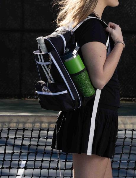 A woman stands courtside in a black tennis outfit, wearing the Fresh Pickle Teddi Pickleball Backpack, which features a roomy interior for essentials like a towel, racquet, water bottle, and ball can. Court lines and net appear in the background.