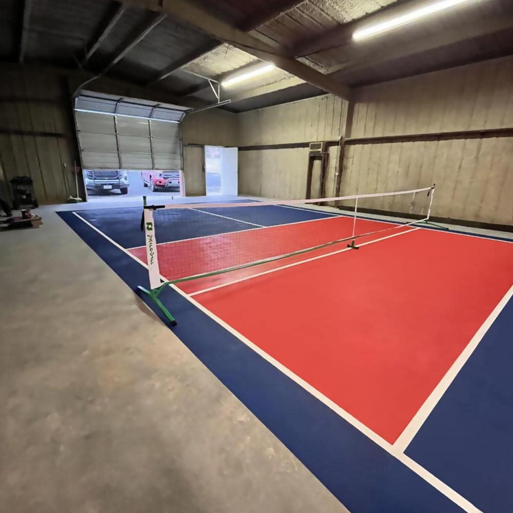 A brightly lit indoor pickleball court painted with SlipDoctors Alpha Grip Non-Slip Stripe and Athletic Court Paint in blue and red, featuring white boundary lines, a white net, and an open rolling door revealing parked cars outside.