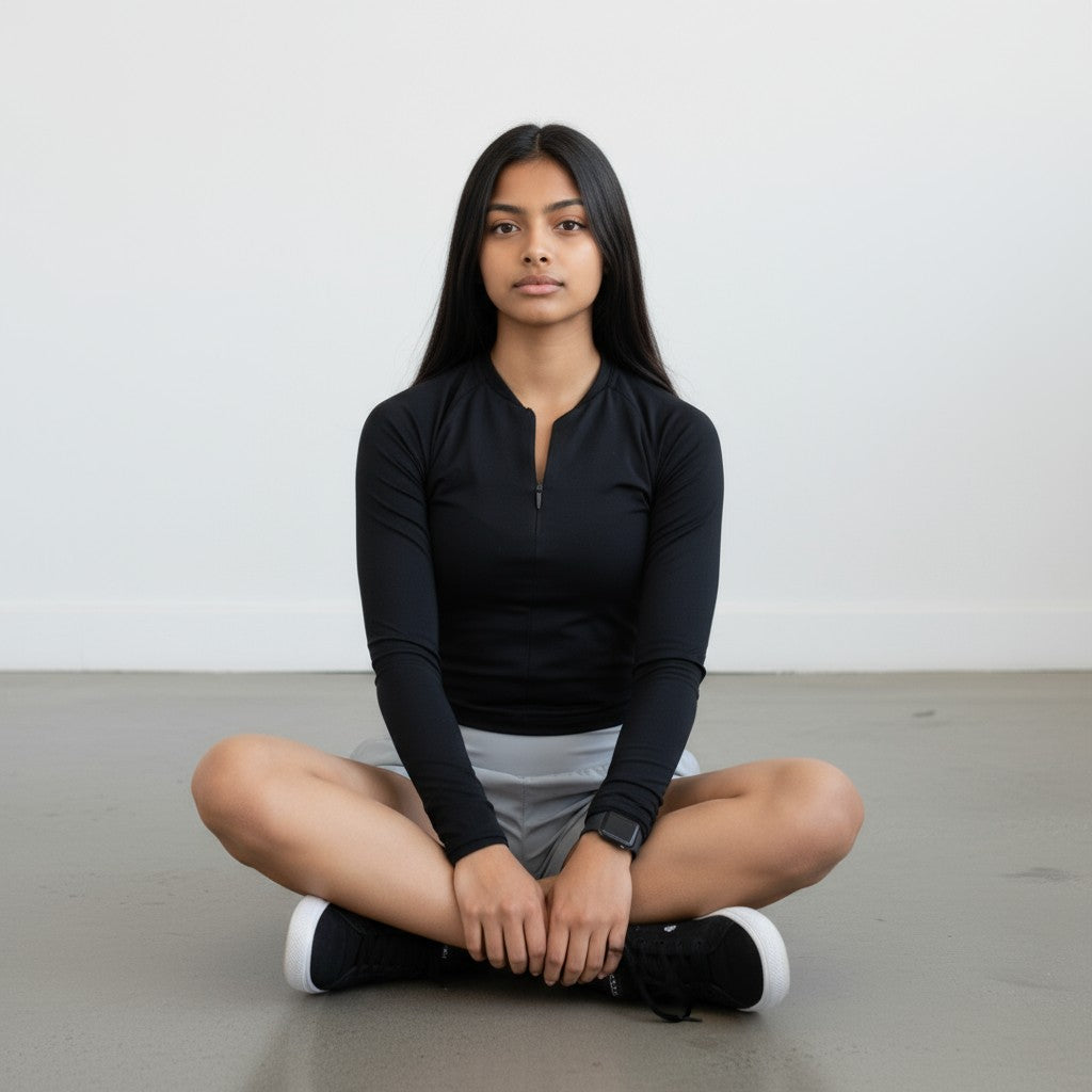 A woman with long dark hair sits cross-legged on the floor in a black DYM Athletics Elite 1/4 Zip Long Sleeve, light gray shorts, and black sneakers against a plain light background, looking forward with a neutral expression.