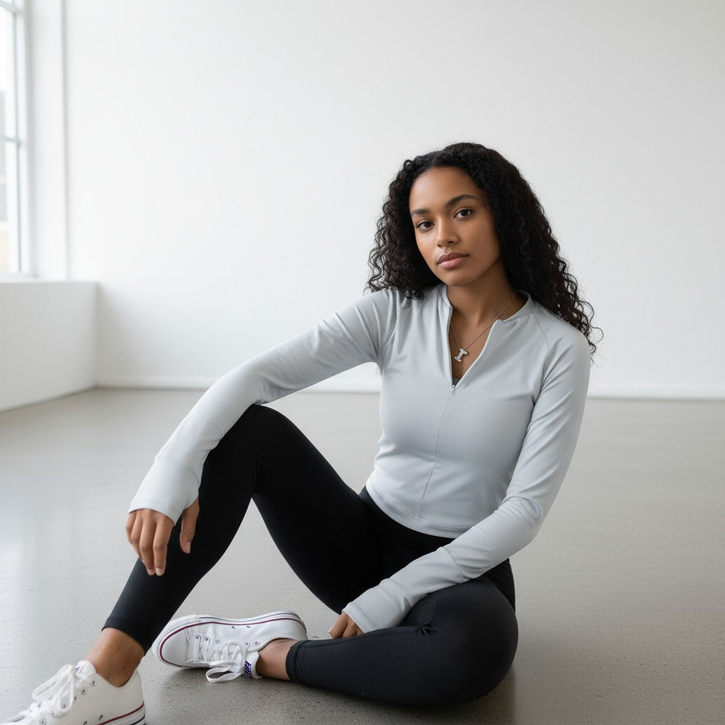 A young woman with long curly hair sits on a light gray floor in a bright, minimalistic room, wearing the DYM Athletics Elite 1/4 Zip Long Sleeve, black leggings, and white sneakers. One leg is bent as she looks calmly at the camera.