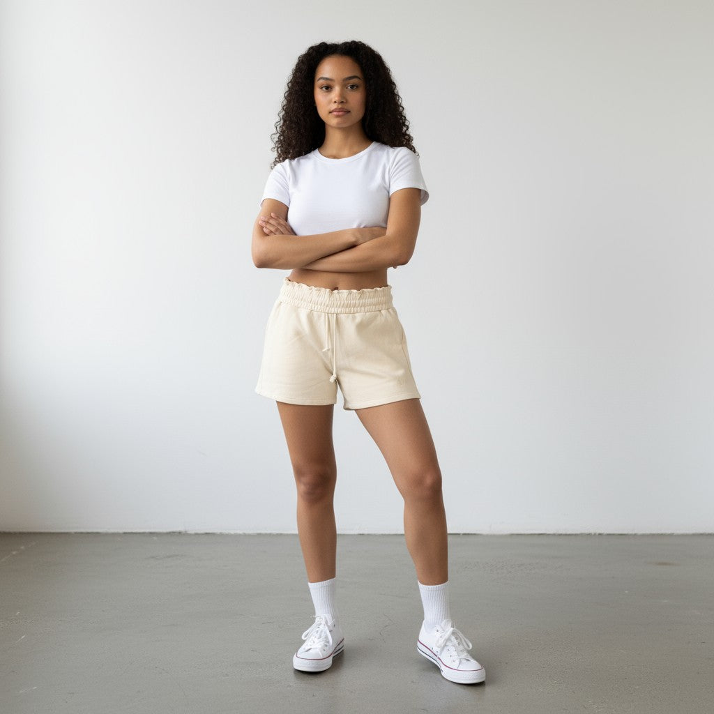 A woman with curly hair stands confidently, wearing DYM Athletics’ Cozy Up Sweat Shorts and a white crop top, paired with crew socks and sneakers on a gray floor against a white wall.