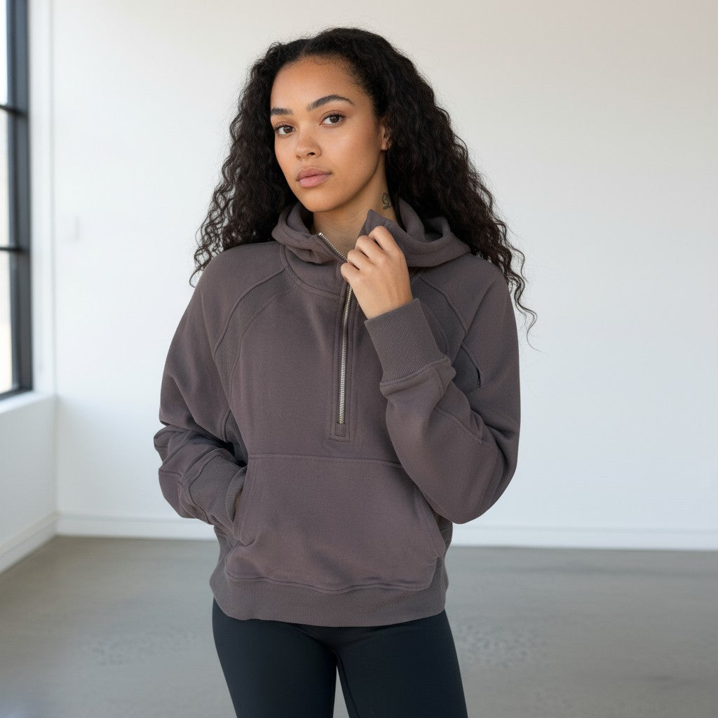 A woman with long curly hair stands indoors in a DYM Athletics Cozy Up Funnel Neck 1/2 Zip Hoodie in dark gray, paired with black leggings. The minimal, bright background highlights her calm expression as her left hand rests near her chin.