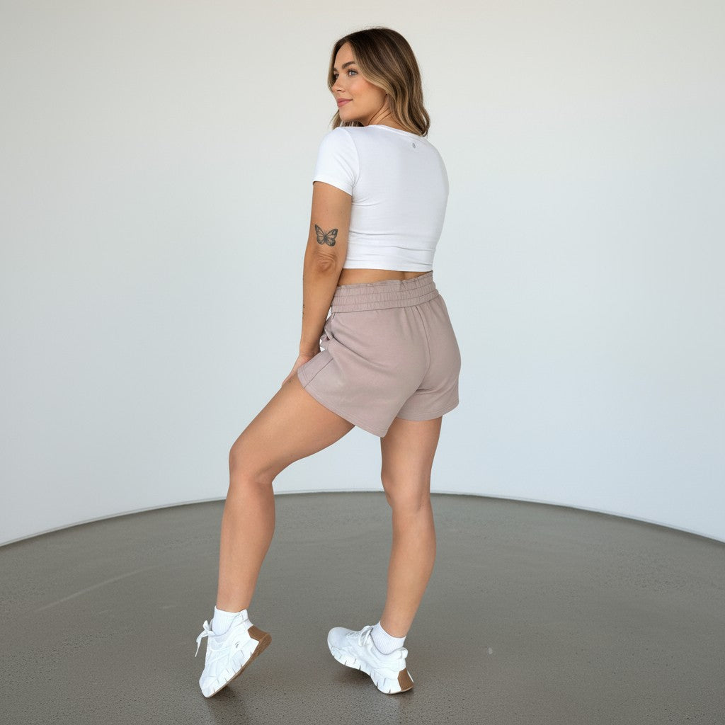 A woman with a butterfly tattoo and wavy, shoulder-length hair stands in a minimal indoor space, facing away but looking over her shoulder. She wears a fitted white crop top, DYM Athletics Cozy Up Sweat Shorts, white crew socks, and sneakers.