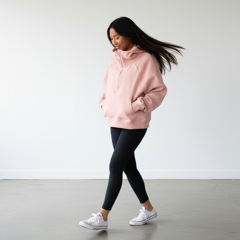A woman with long dark hair wears the DYM Athletics Cozy Up Funnel Neck 1/2 zip Hoodie in pink, paired with black leggings and white sneakers. Walking indoors on a gray floor, she has her hands in her hoodie pocket and looks down, relaxed.