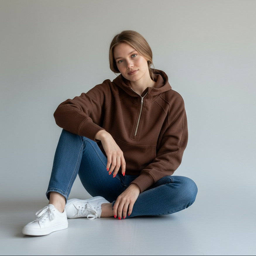 A young woman with light brown hair sits on the floor against a plain light background, looking relaxed with crossed legs. She wears blue jeans, white sneakers, and the Cozy Up Funnel Neck 1/2 Zip Hoodie by DYM Athletics.
