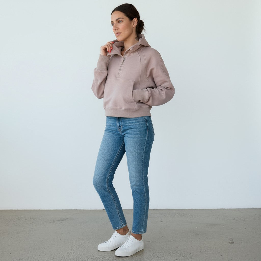 A woman stands against a white wall, wearing the DYM Athletics Cozy Up Funnel Neck 1/2 Zip Hoodie with blue jeans and white sneakers. Her hair is pulled back, one hand in her pocket as she thoughtfully touches her chin.