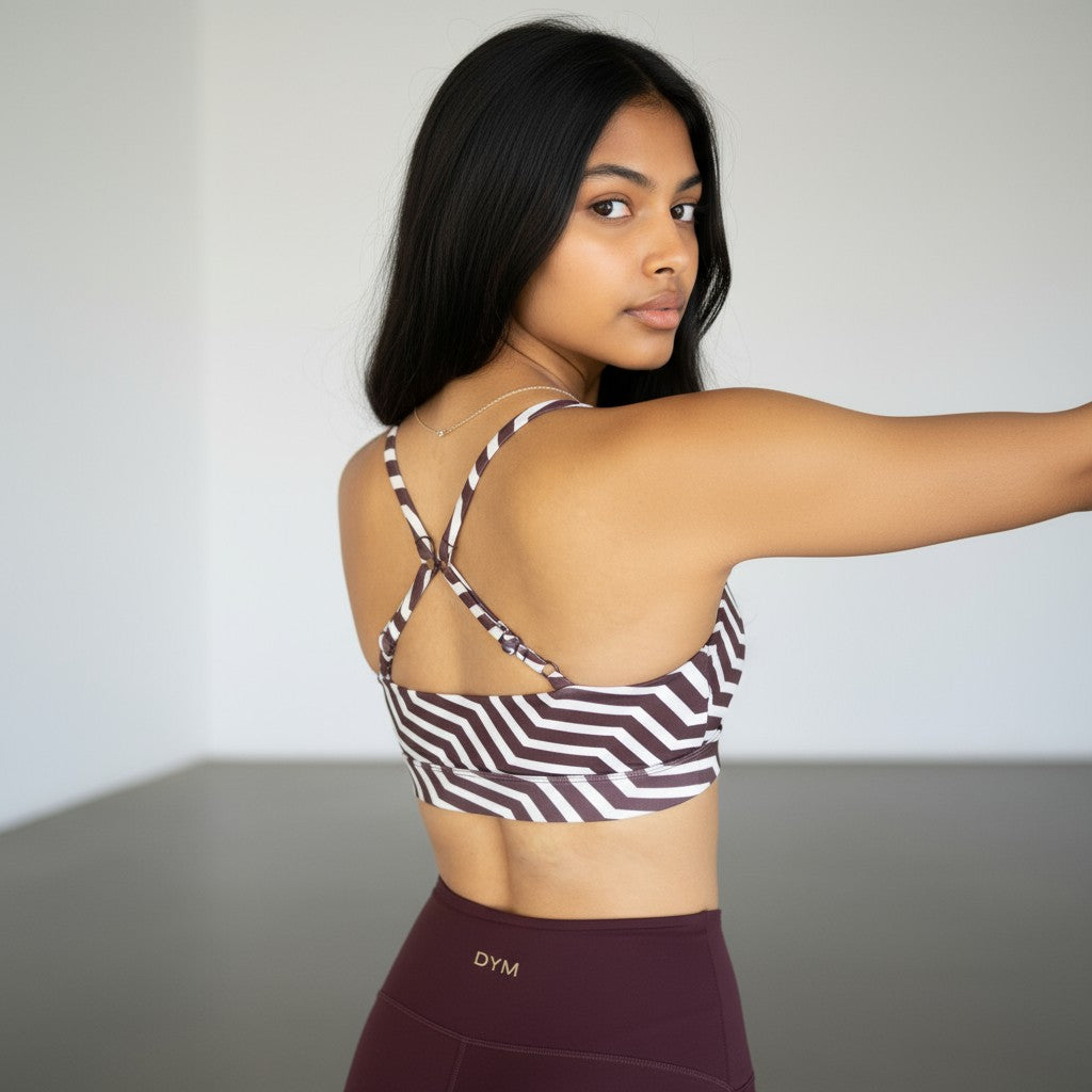 A woman with long dark hair wears a striped DYM Athletics Trainer Performance 2-Way Bra (A-D cup, medium-high support) with crisscross back and maroon leggings featuring a DYM logo on the waistband, posing in a bright, minimalist room.