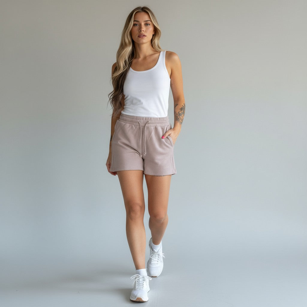 A woman with long, wavy blonde hair wears a white tank top, DYM Athletics Cozy Up Sweat Shorts in light mauve, and white sneakers. She stands against a plain light gray background with one hand in her pocket, looking forward neutrally.