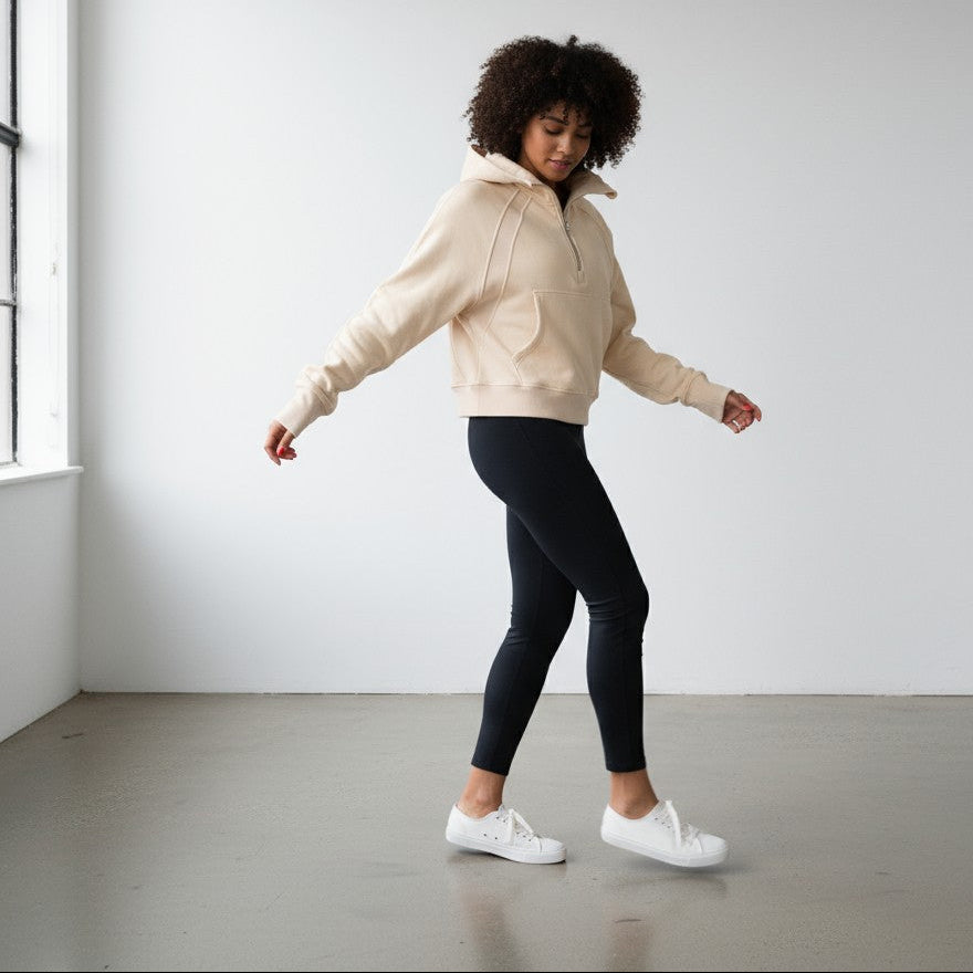 A woman with curly hair wears the DYM Athletics Cozy Up Funnel Neck 1/2 Zip Hoodie, black leggings, and white sneakers as she stands indoors on a gray floor near a large window, turning gracefully with eyes closed against a plain white wall.