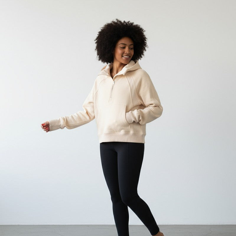 A woman with curly hair smiles in a minimalist room, wearing the DYM Athletics Cozy Up Funnel Neck 1/2 Zip Hoodie with front pockets and black leggings. She looks relaxed and happy, her left arm slightly extended as she looks away from the camera.