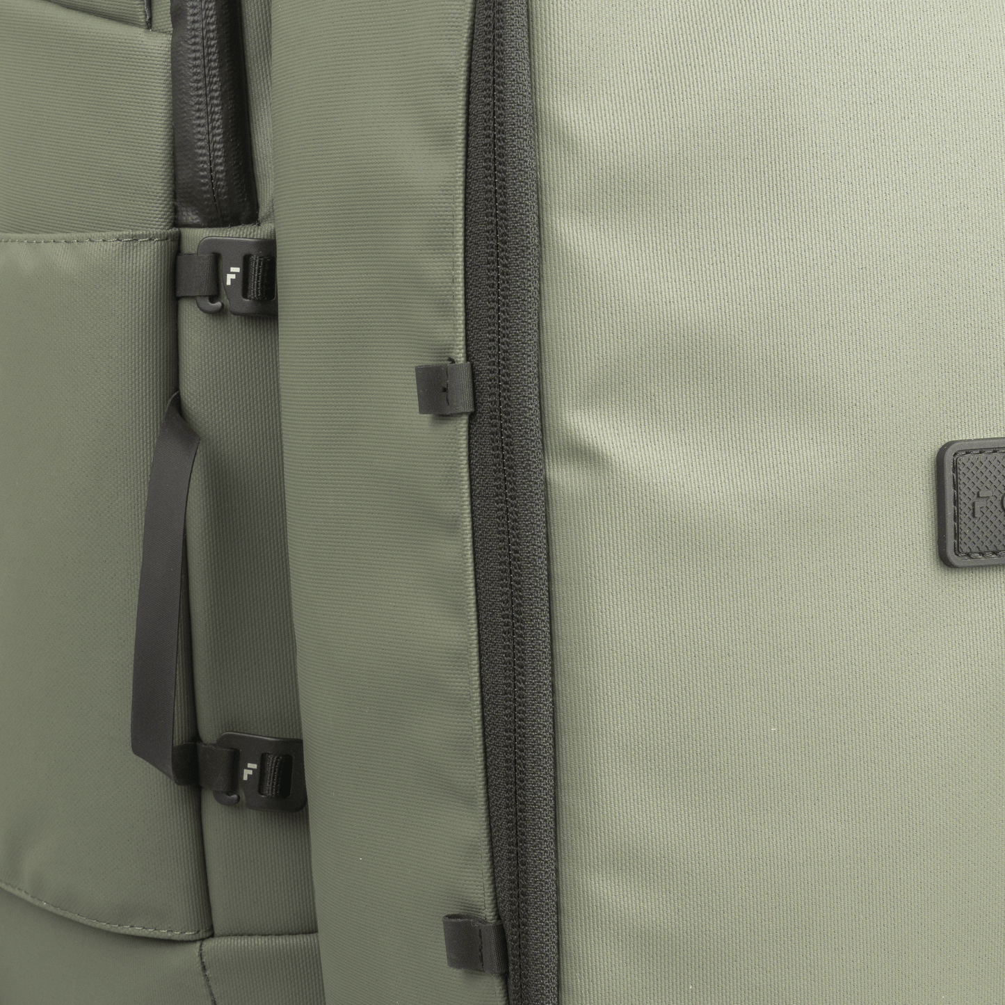 Close-up of a green backpack with black vertical zipper, buckles, and straps. Its durable fabric pairs well with the FORWRD Court Caddy Shoe Cube. A small square black patch with embossed text sits on the right side.