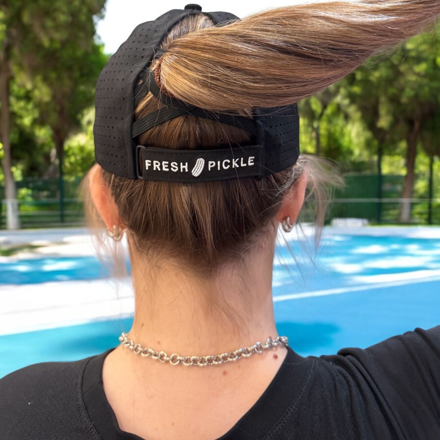 A blonde-haired person in a ponytail sports a Fresh Pickle Pickleball Cap on a blue pickleball court, accessorized with silver hoop earrings and a chain necklace, with trees and a green fence behind them.