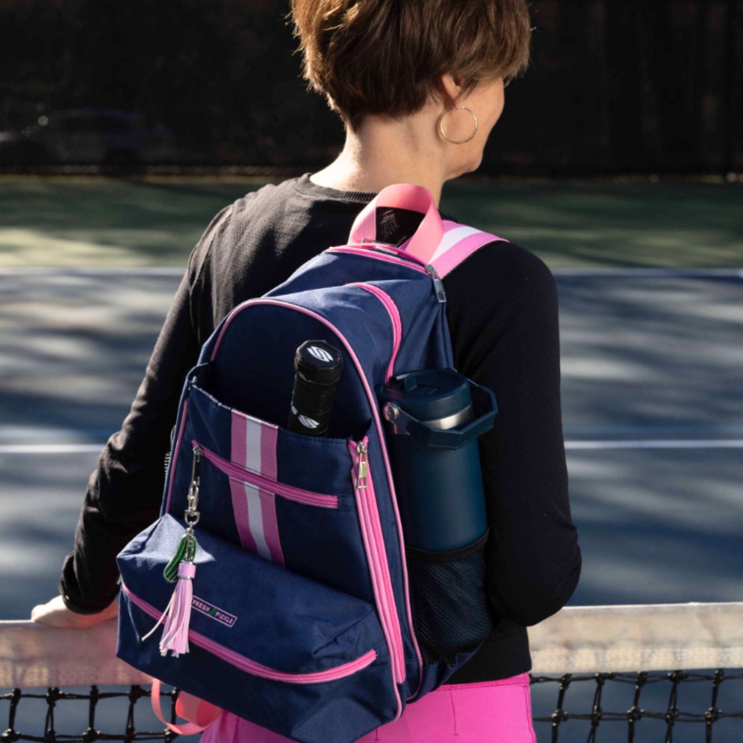 A woman with short brown hair stands by a tennis court, seen from behind. She wears a black top, pink skirt, and carries a navy blue backpack holding a Fresh Pickle Tumbler by Fresh Pickle and a tennis ball canister.