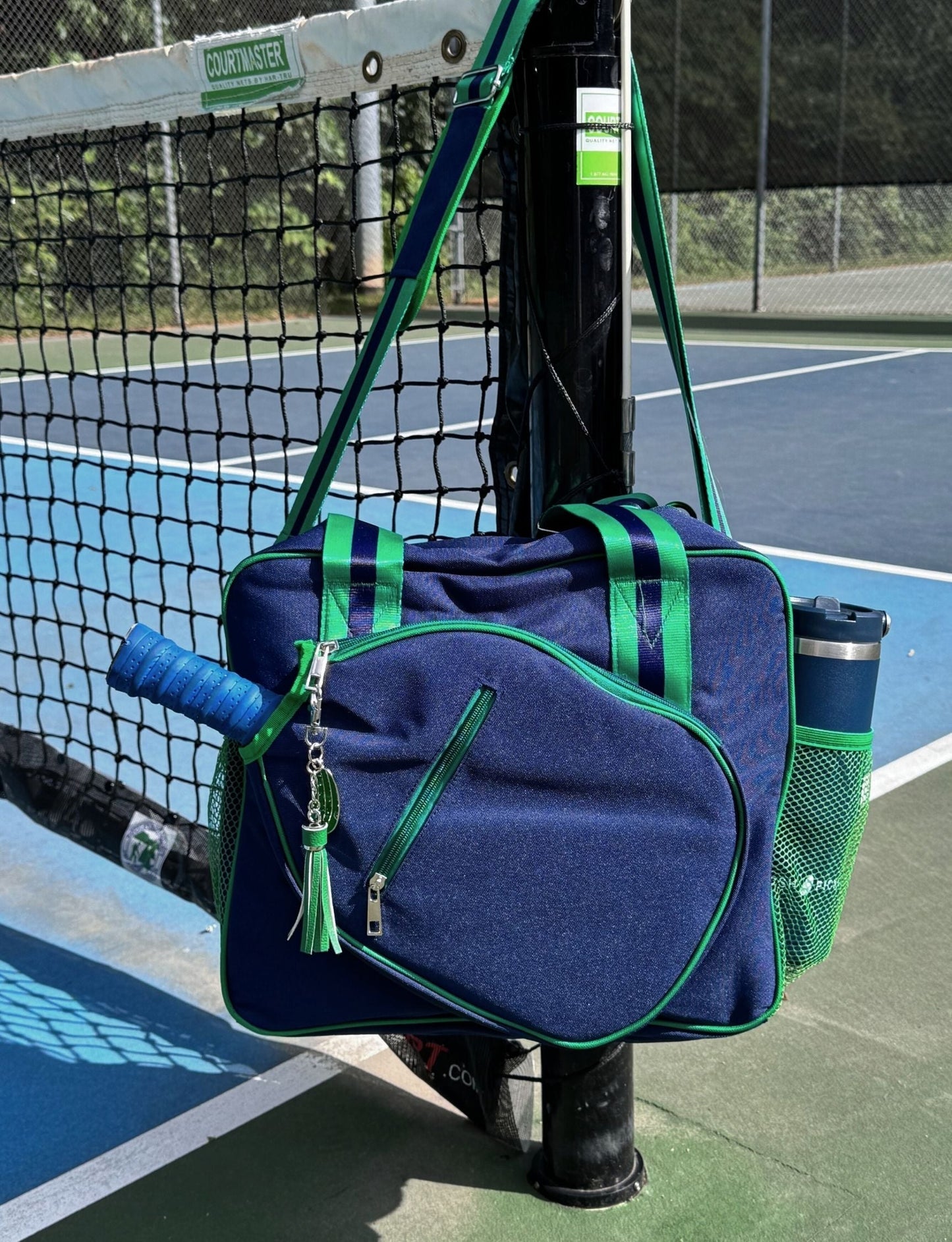 The Lola Pickleball Tote Bag by Fresh Pickle, featuring navy blue with green trim, hangs on a tennis net post. A blue-handled racket and keychain are attached, while the green mesh side pocket holds a blue bottle. The court is visible behind.
