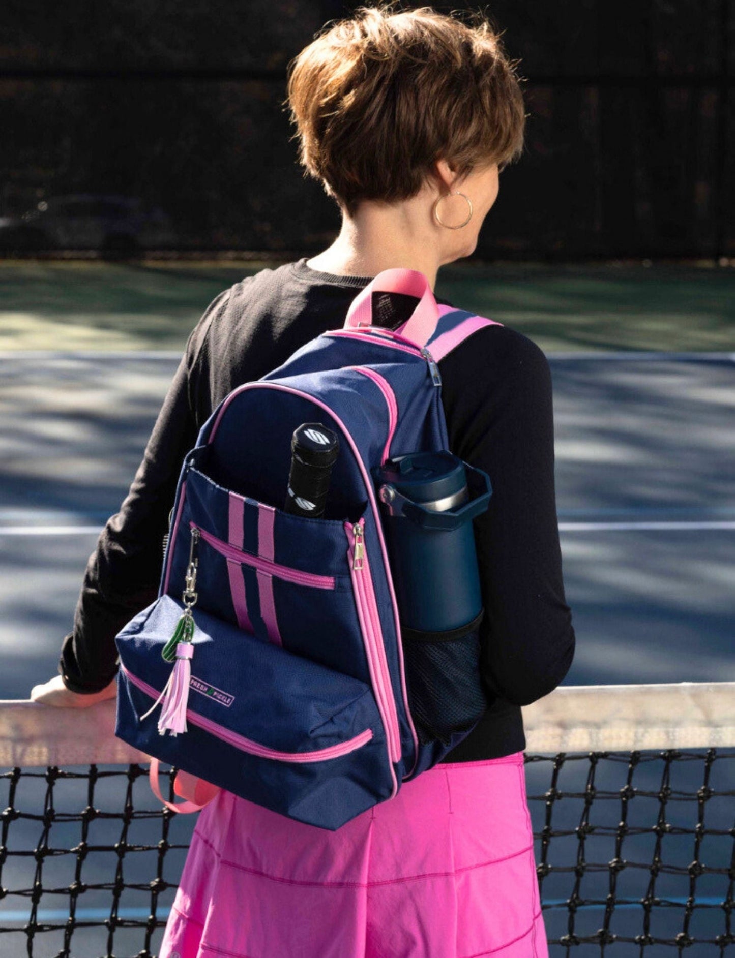 A person with short brown hair, in a black top and pink tennis skirt, stands by a net. They're carrying the Fresh Pickle Teddi Pickleball Backpack, which holds a water bottle and can, on a court with a shadowy background.