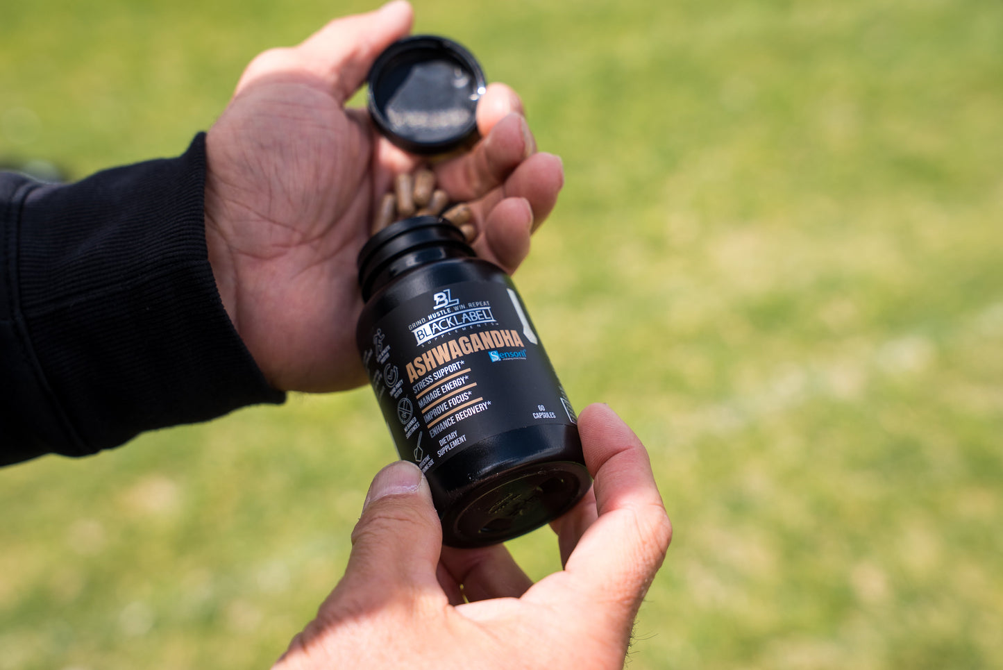 A person holds an open bottle of BLACKLABEL Supplements SENSORIL® ASHWAGANDHA capsules outdoors, with a grassy blurred background. The label highlights ashwagandha and black pepper extract for optimal absorption.