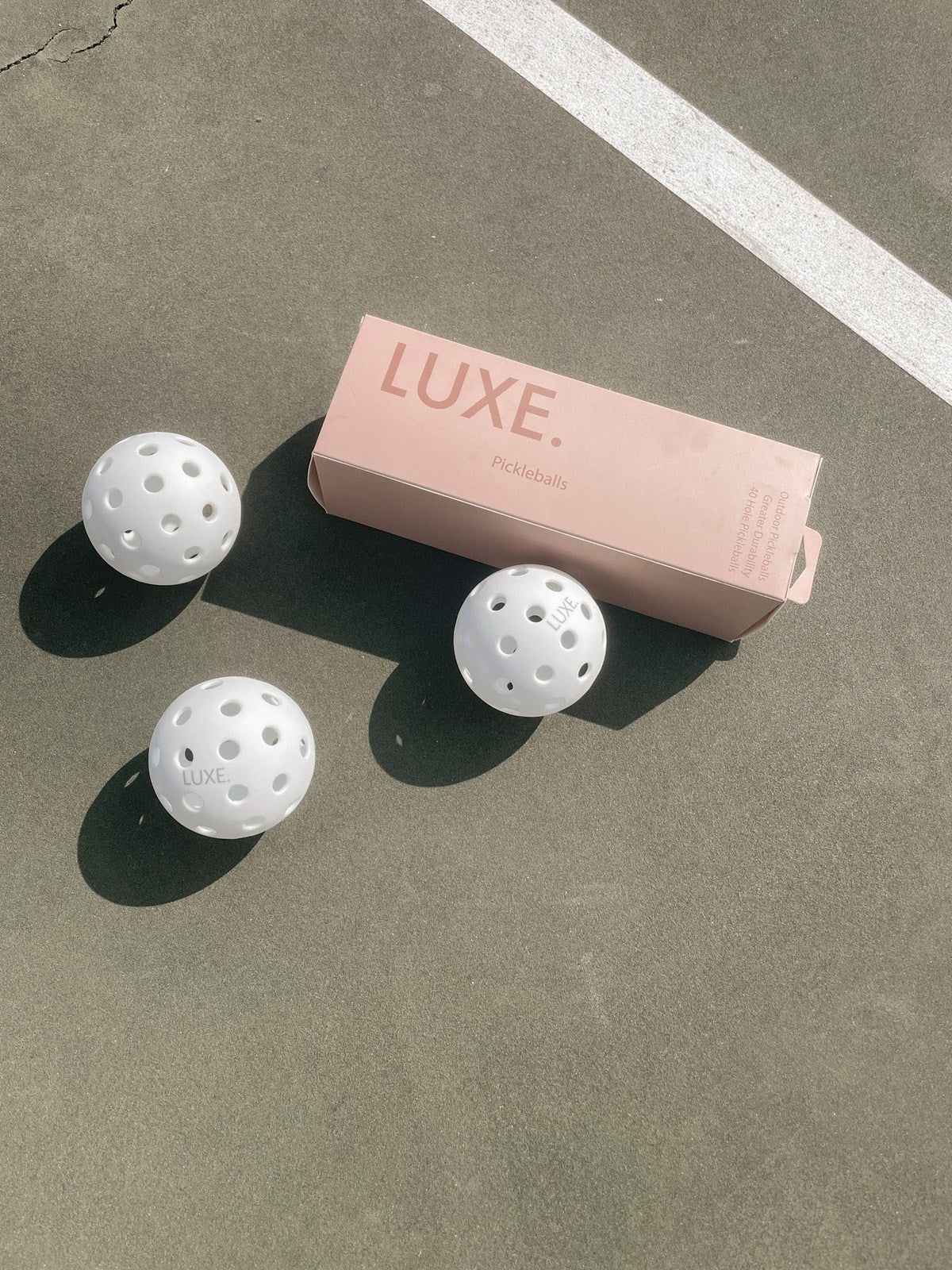 Three LUXE Pickleball White Pickleballs (pack of 3, each with 40 holes) sit on a sunlit outdoor court beside a rectangular pink LUXE Pickleball box, their shadows crossing a green court marked by a diagonal white line.