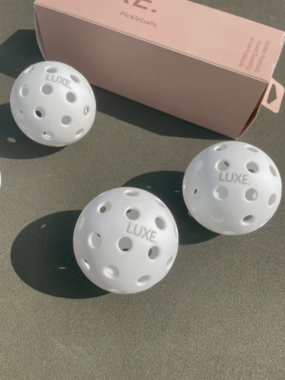 Three LUXE Pickleball White Pickleballs, each featuring multiple holes, sit on a smooth surface in front of a light pink box labeled "Pickleballs," with natural sunlight casting soft shadows across the scene. Pack contains 3 pickleballs.