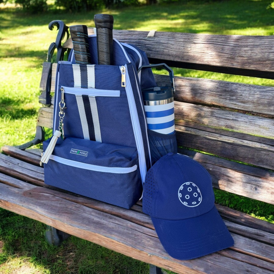 The Fresh Pickle Teddi Pickleball Backpack, in blue, sits on a park bench holding two paddles, a striped water bottle, and a pickleball-themed cap. Sunlight and green grass brighten the scene.