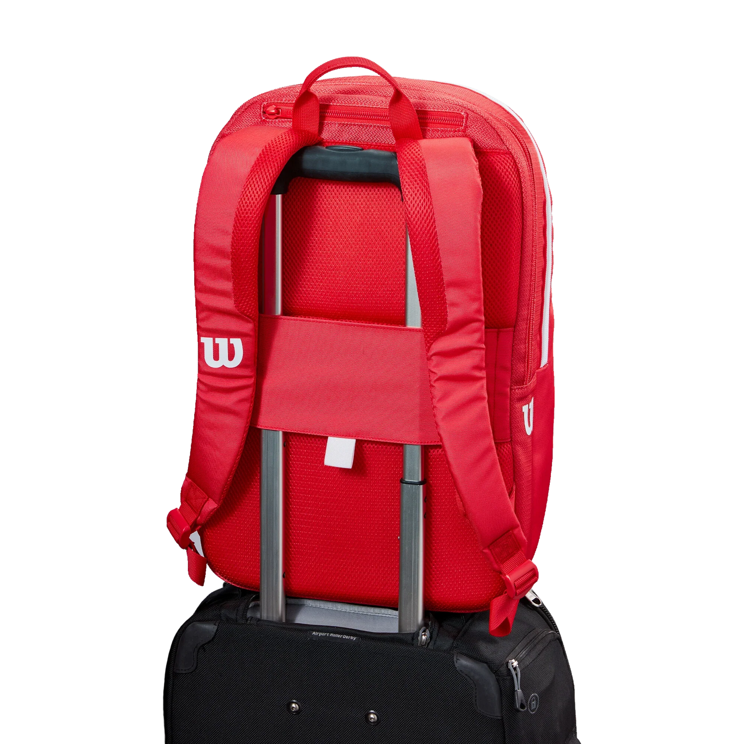The Wilson Tour Red Padel Backpack from Padel USA, featuring padded straps and a white W logo, is attached to the handle of a black rolling suitcase via its convenient luggage sleeve. The background is plain white.