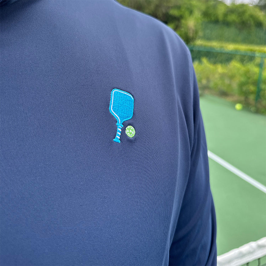 A close-up of a person wearing the Swinton Pickleball Court Performance Quarter Zip Pullover in Navy, featuring an embroidered blue paddle and green pickleball on the chest, with a green court and greenery in the background.