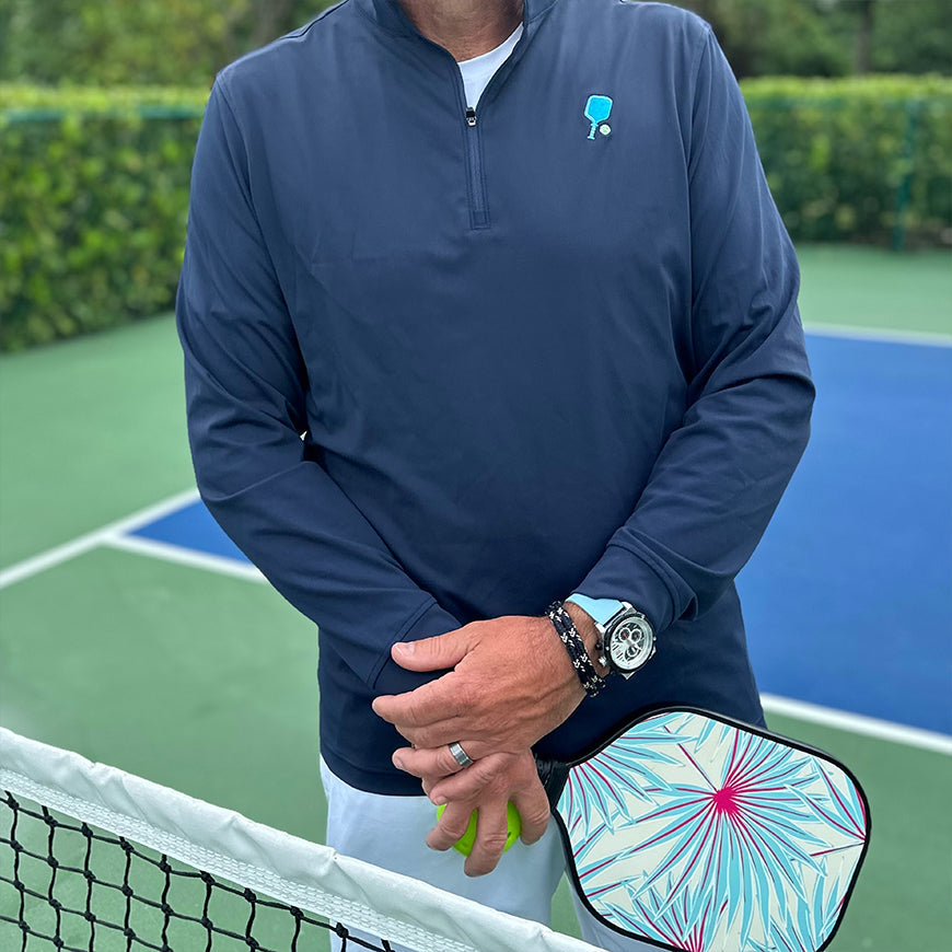 On a tennis court, a person stands in front of the net with crossed arms, wearing the Swinton Pickleball Court Performance Quarter Zip Pullover in Navy and holding a colorful pickleball paddle. Greenery is visible in the background.