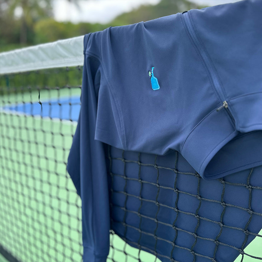 The Swinton Pickleball Court Performance Quarter Zip Pullover in Navy, featuring a subtle embroidered logo, hangs over the black net of an outdoor tennis court; the out-of-focus background shows the court and green trees.