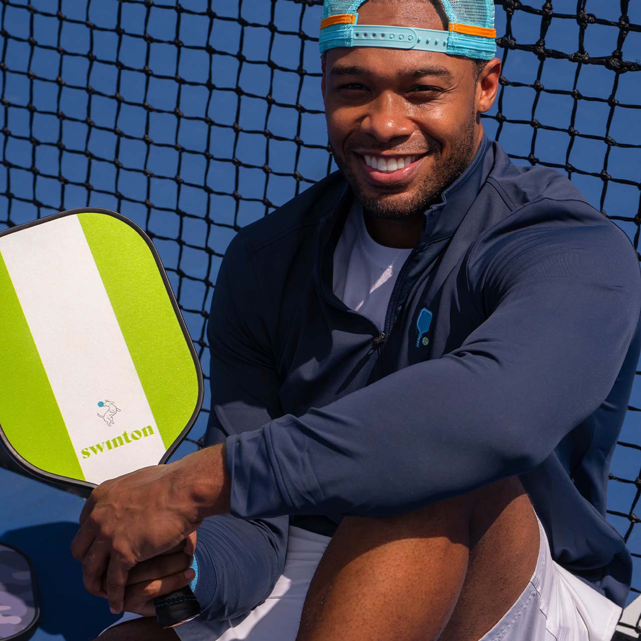 A man smiles while sitting on an outdoor pickleball court, wearing a Swinton Pickleball Court Performance Quarter Zip Pullover in Navy and holding a bright green and white Swinton paddle. The net and blue court are visible behind him.