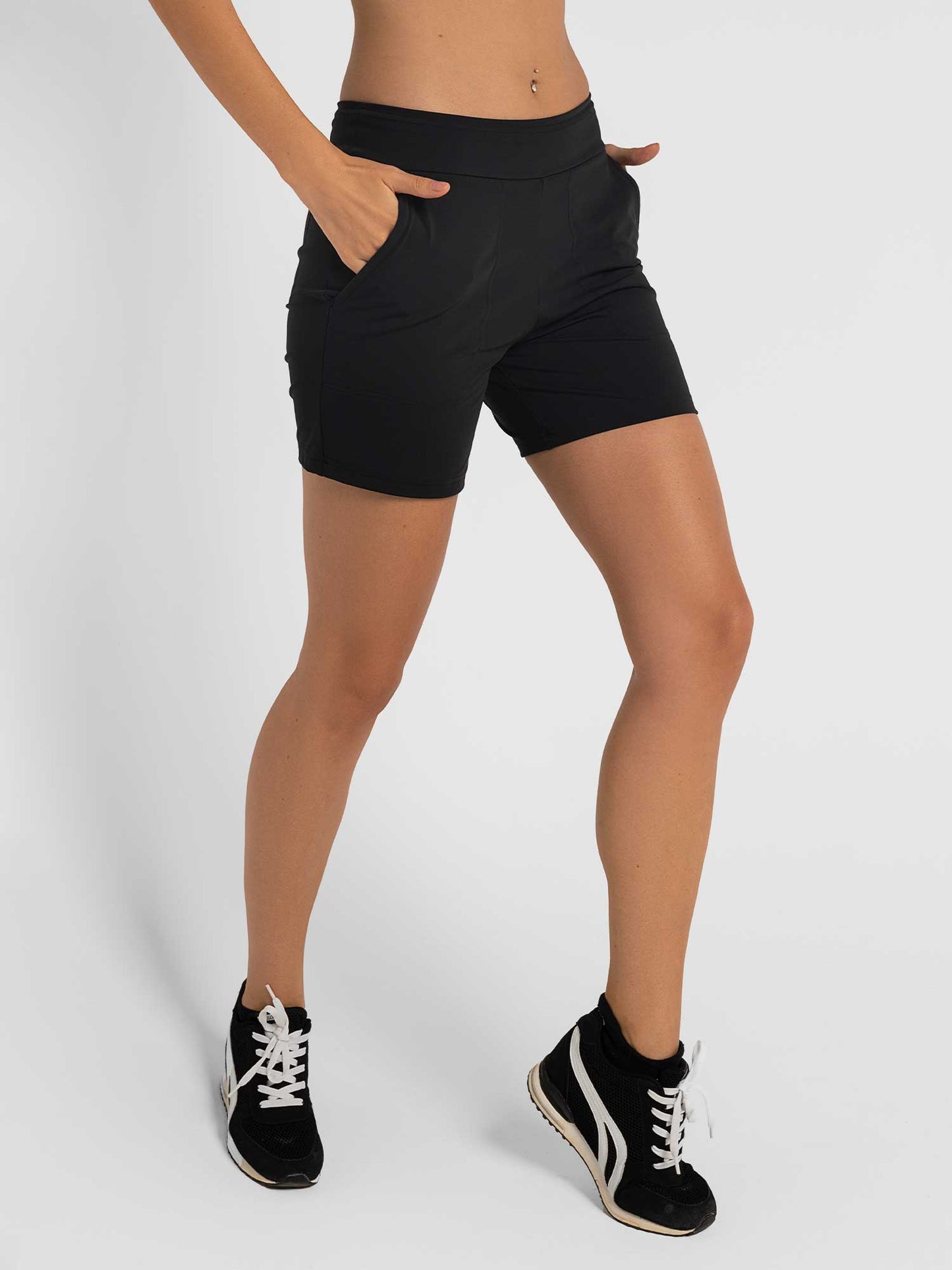 A person models the inPhorm NYC Women's Cleo Short—stylish and functional for active lifestyles—in black, paired with black and white sneakers. The plain white background highlights its moisture management for all-day comfort.