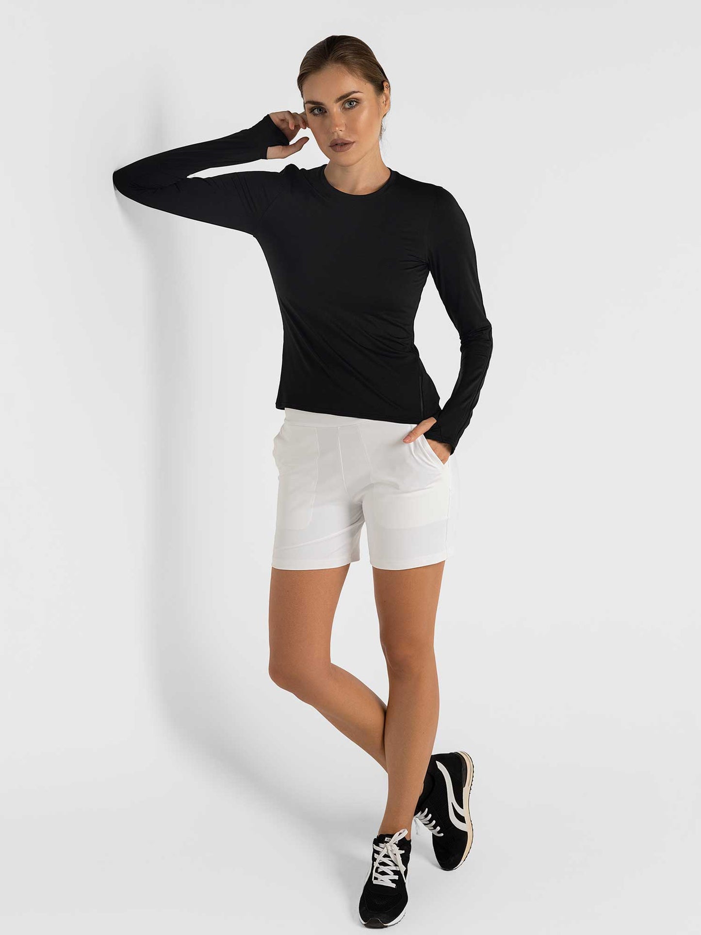 A woman stands against a white wall in a black long-sleeve shirt, inPhorm NYC Women's Cleo Short - Stylish & Functional for Active Lifestyles in white, with pockets, paired with black athletic shoes. She rests her left elbow on the wall and looks ahead.